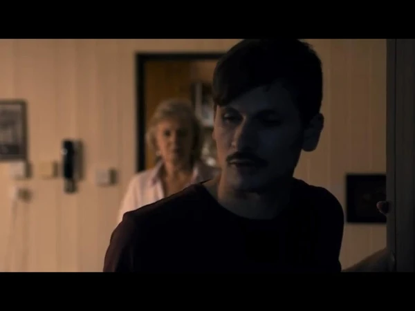 A young man stands in a doorway with an older woman in the background in "Saudade".